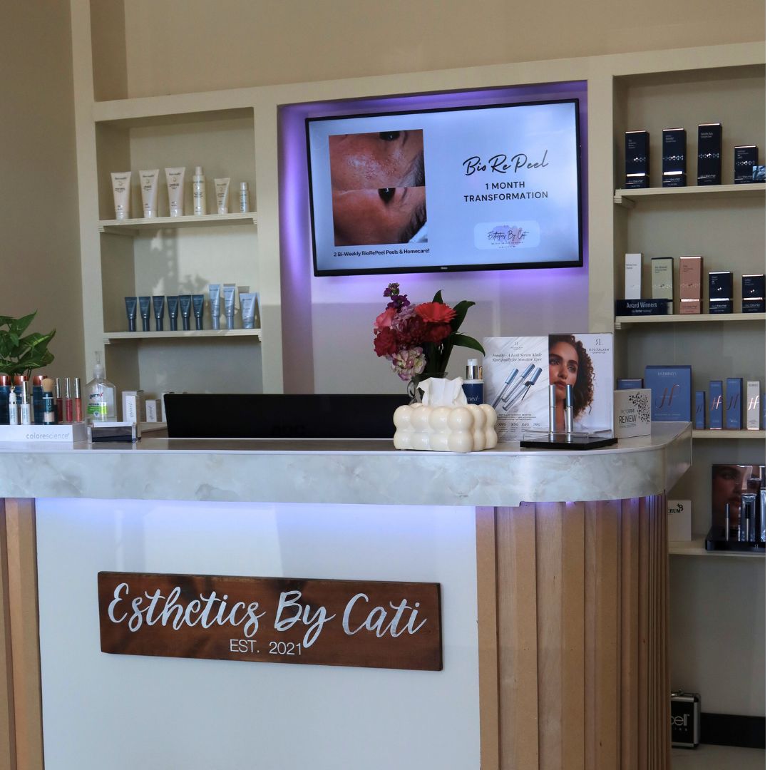 Front desk of a skincare clinic named 'Esthetics By Cati' with a monitor displaying a skincare transformation.